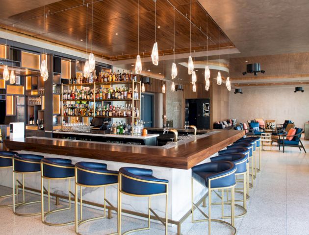 This image shows a modern bar with high chairs, a large liquor selection, unique hanging lights, and a comfortable seating area in the back.