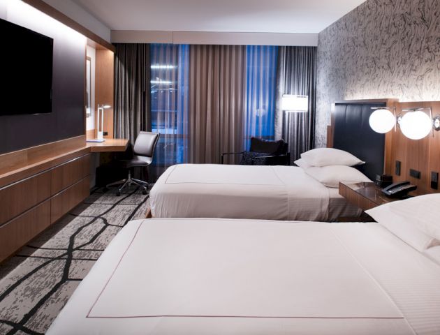 The image shows a modern hotel room with two neatly made beds, a flat-screen TV, a desk, and floor-to-ceiling windows with curtains.
