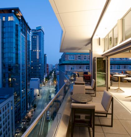 A rooftop terrace at dusk with tables and seating, overlooking a cityscape with tall buildings and illuminated streets.