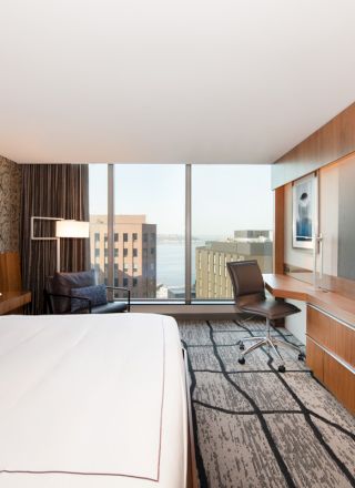 A modern hotel room with a large bed, wall-mounted TV, work desk with chair, armchair, and window with a cityscape view.