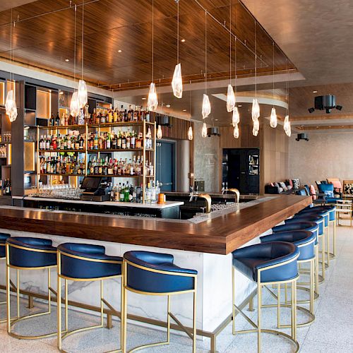 A modern bar with blue barstools, a well-stocked shelf of drinks, pendant lights, wood accents, and a lounge area in the background.