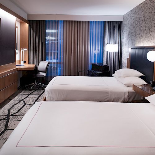 The image shows a modern hotel room with two beds, a wall-mounted TV, a desk with a chair, and stylish decor and lighting.