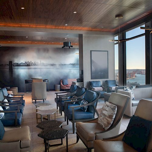 A modern lounge with ample seating, large windows overlooking water and a ferris wheel, and a cityscape mural on the back wall.