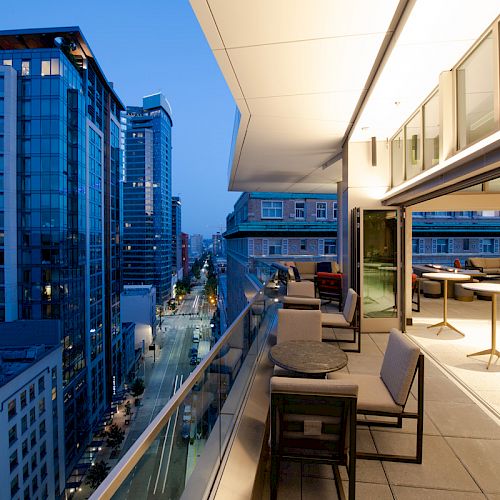 This image features a modern urban balcony with seating, overlooking city buildings at dusk. The interior is well-lit, contrasting with the evening sky.
