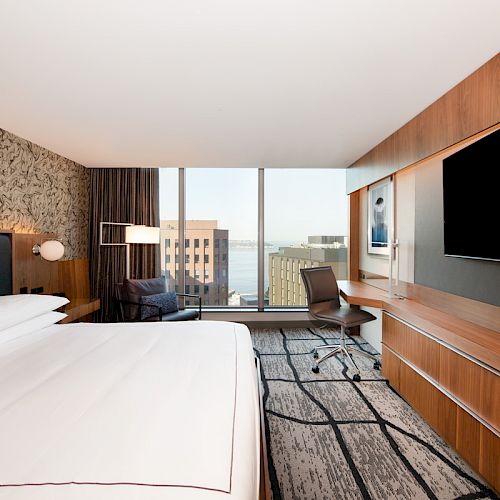 Modern hotel room with a large bed, wall-mounted TV, desk, chair, floor-to-ceiling windows with city view, and contemporary decor.
