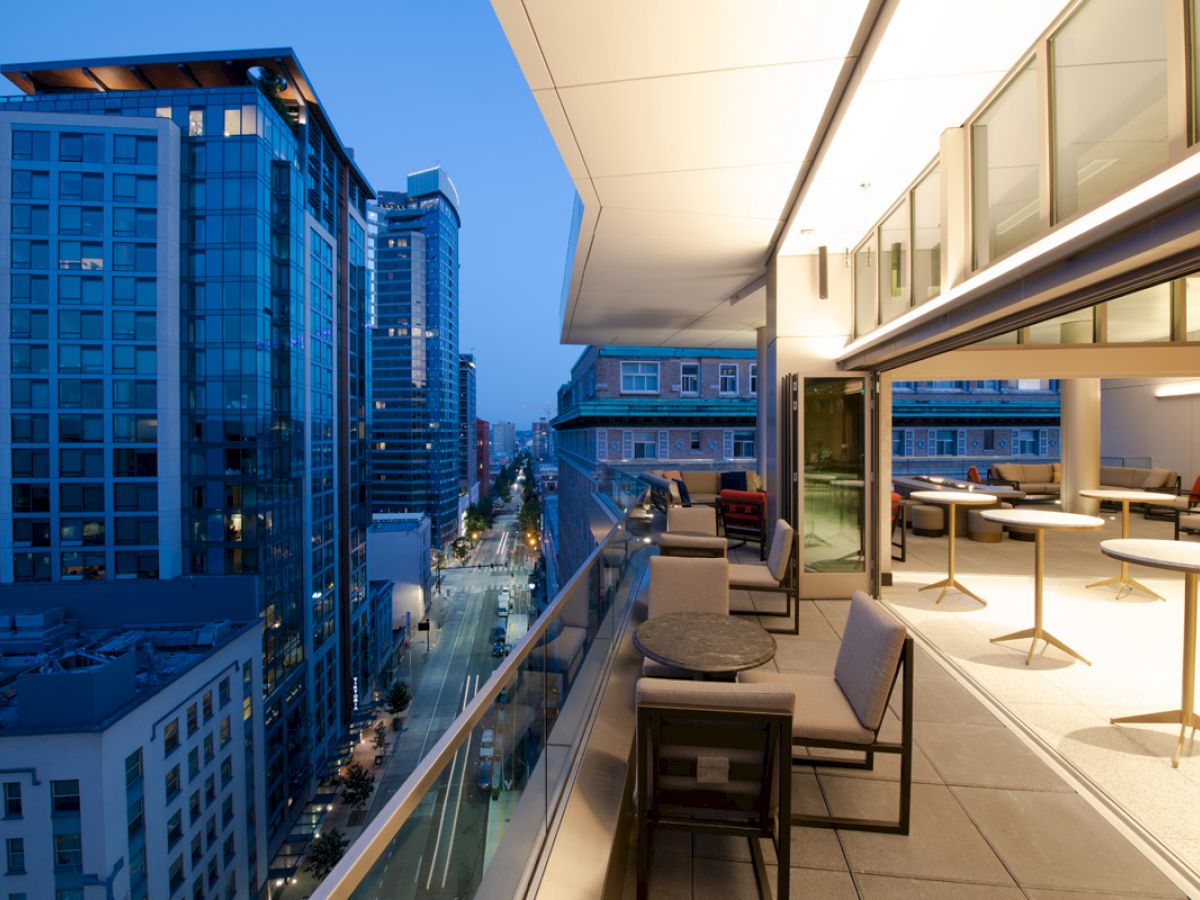 A modern balcony overlooks a cityscape with tall buildings and streetlights, featuring outdoor seating and tables under warm lighting.