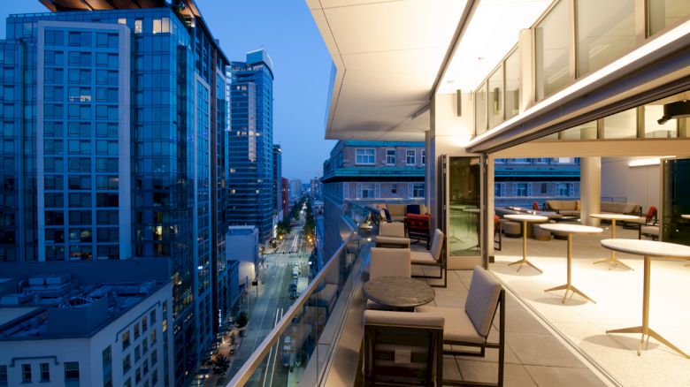 A modern balcony overlooks a cityscape with tall buildings and streetlights, featuring outdoor seating and tables under warm lighting.