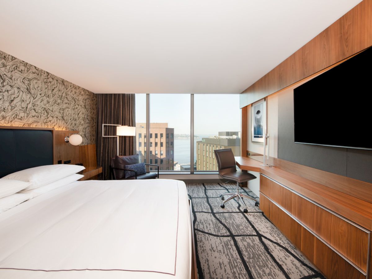 A modern hotel room with a large bed, a desk, a chair, a flat-screen TV, and a large window offering a city view.