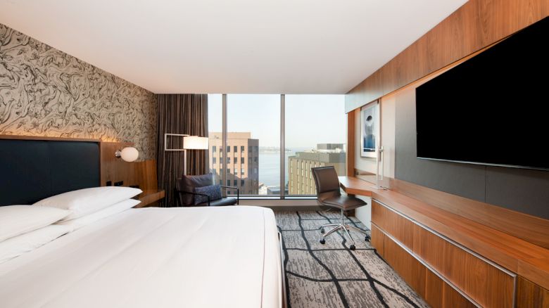 A modern hotel room with a large bed, a desk, a chair, a flat-screen TV, and a large window offering a city view.