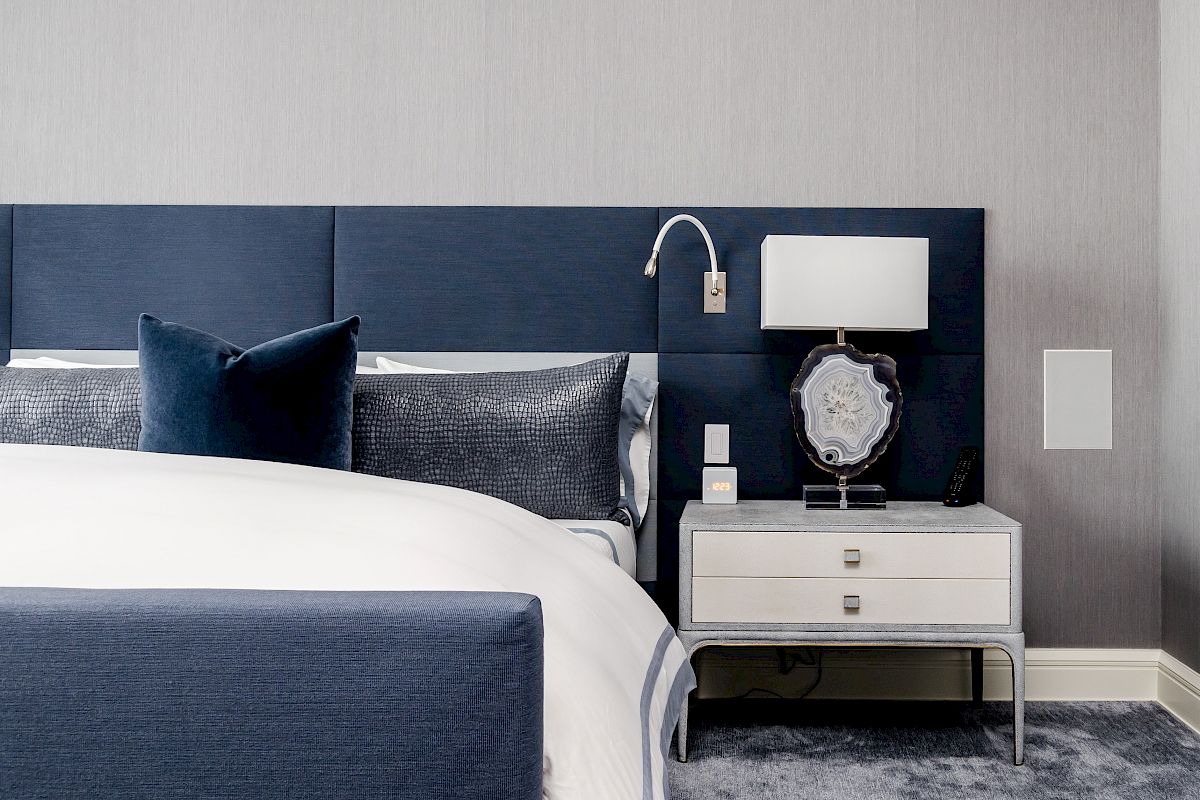 A modern bedroom with a blue and white color scheme, featuring a bed with blue pillows, nightstand, lamp, and a wall-mounted reading light.