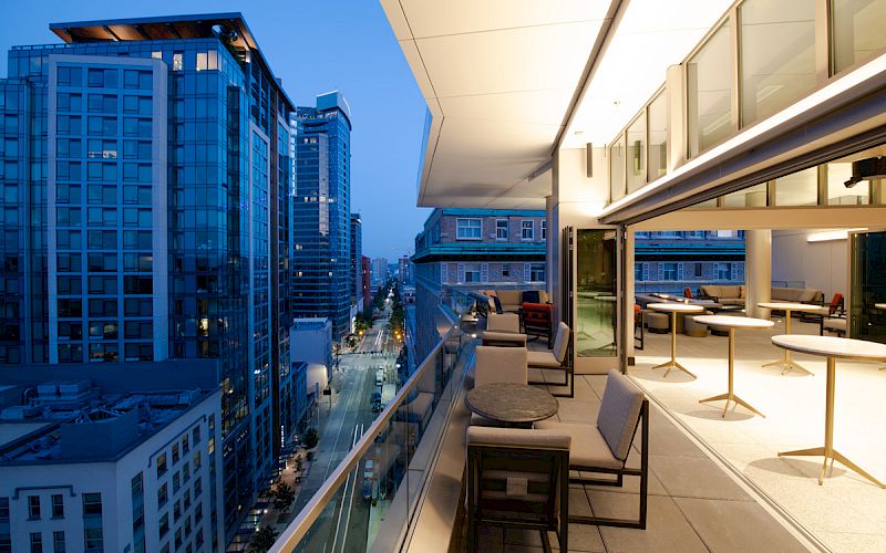 The image shows a modern balcony with tables and chairs overlooking a cityscape of tall buildings during dusk, featuring a serene evening ambiance.