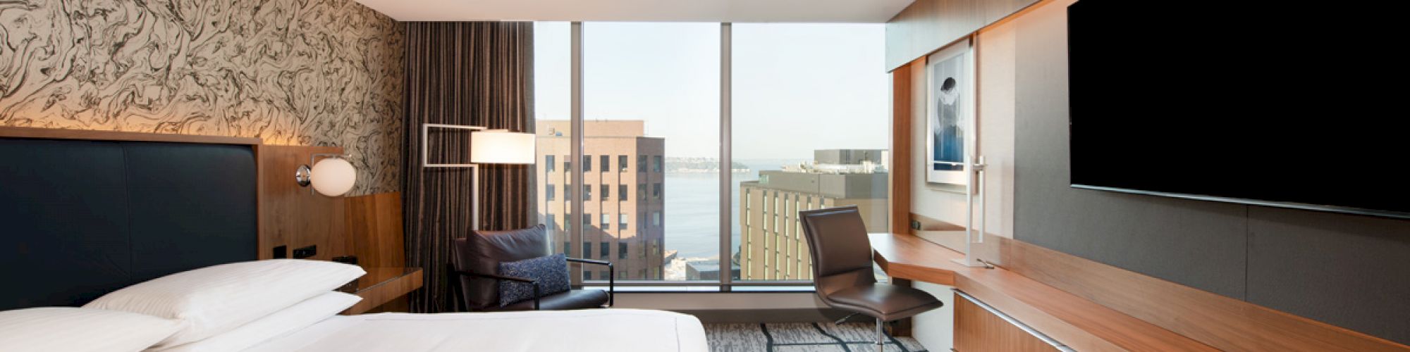A modern hotel room with a large bed, a wall-mounted TV, a desk with a chair, and a window view of city buildings and water in the distance.