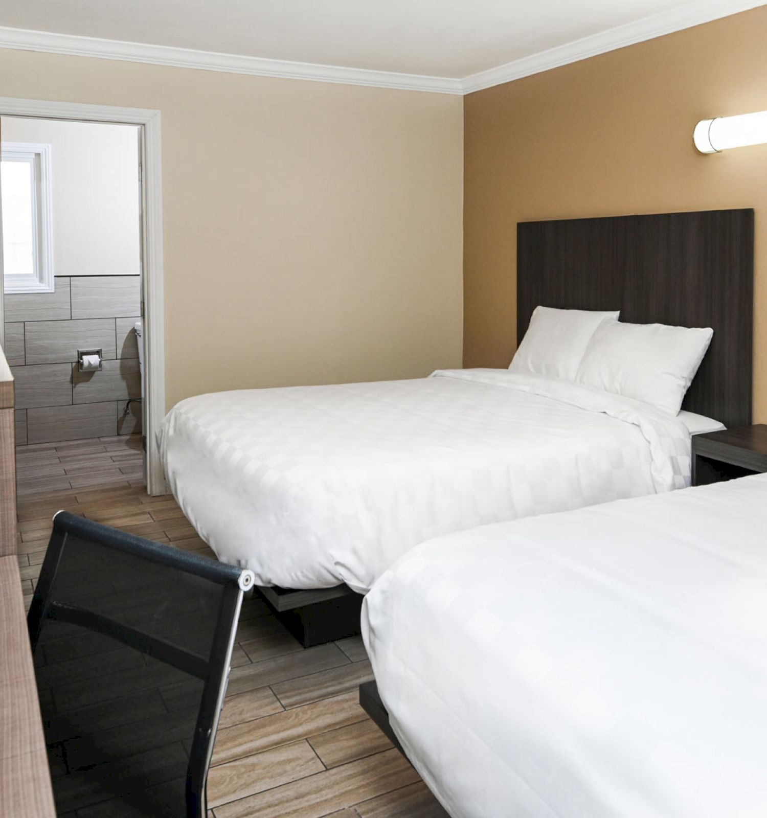 The image shows a hotel room with two beds, a desk, a flat-screen TV, and an adjoining bathroom. The room has a modern and clean design.