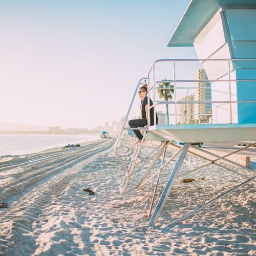 A person is sitting on a lifeguard tower on a sandy beach, with buildings and palm trees in the background. The beach appears calm and peaceful.