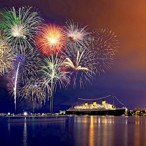 A night scene with colorful fireworks exploding in the sky over a large illuminated ship docked by the waterfront.