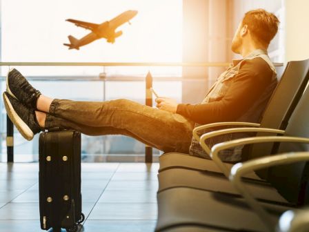 A person is sitting in an airport waiting area with feet propped on luggage, watching an airplane take off through the large window.