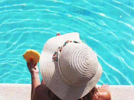 A person wearing a sunhat and a swimsuit sits by a pool, holding what appears to be a slice of fruit, with their feet dangling above the water.